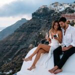 Coppia elegante seduta su un muretto bianco a Santorini al tramonto che guarda insieme lo schermo di un telefono con due calici in mano